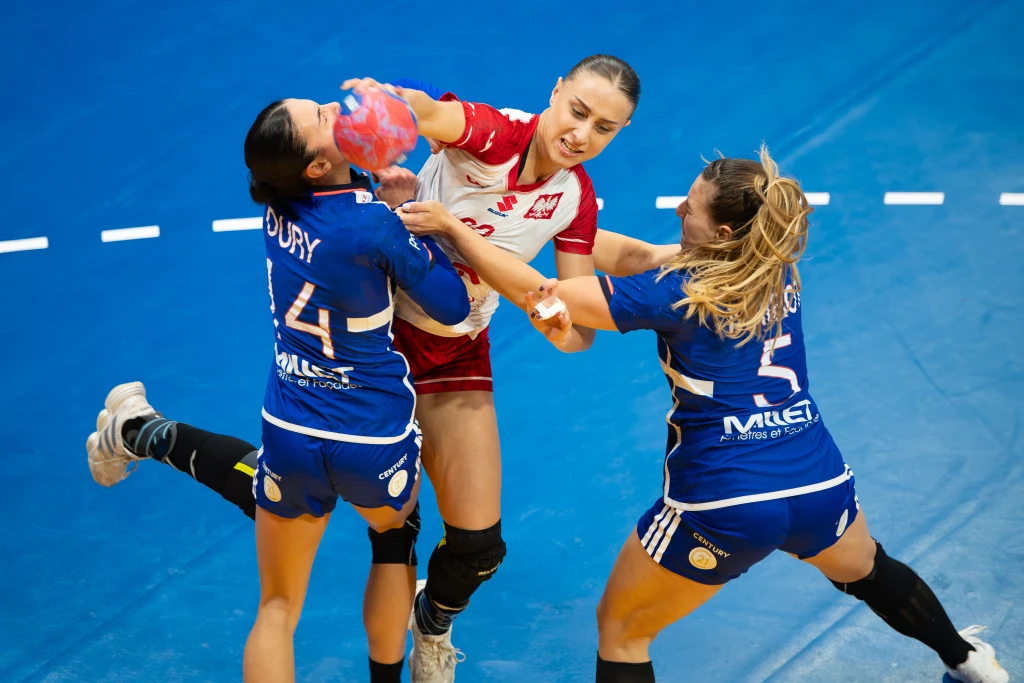 Dynamiczna akcja podczas meczu piłki ręcznej kobiet, zawodniczka w biało-czerwonym stroju walczy o piłkę z dwiema zawodniczkami drużyny przeciwnej w niebieskich strojach, na tle niebieskiej podłogi hali sportowej.