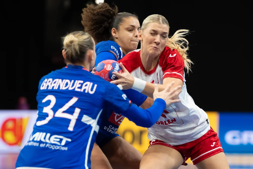 Dwie zawodniczki walczą o piłkę podczas meczu piłki ręcznej, jedna ubrana w niebieski strój z numerem 34, druga w biało-czerwonym stroju, w tle widoczna jeszcze jedna zawodniczka i rozmyte elementy boiska.