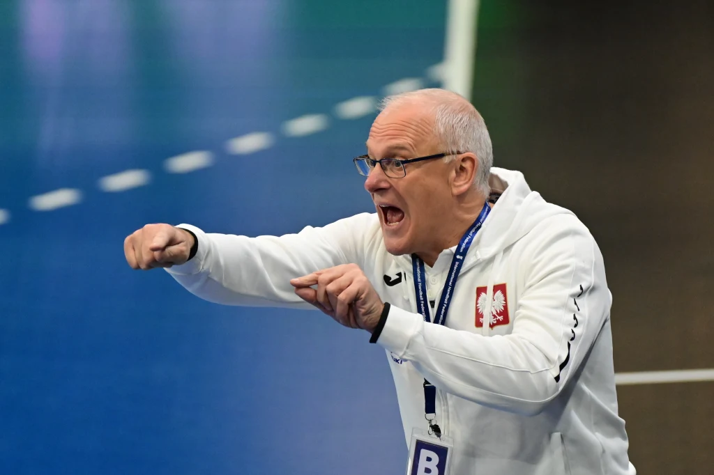 Mężczyzna w białej bluzy z godłem Polski energicznie gestykuluje, prawdopodobnie wydając polecenia podczas meczu sportowego, z wyrazem silnych emocji na twarzy oraz zawieszką identyfikacyjną na szyi.