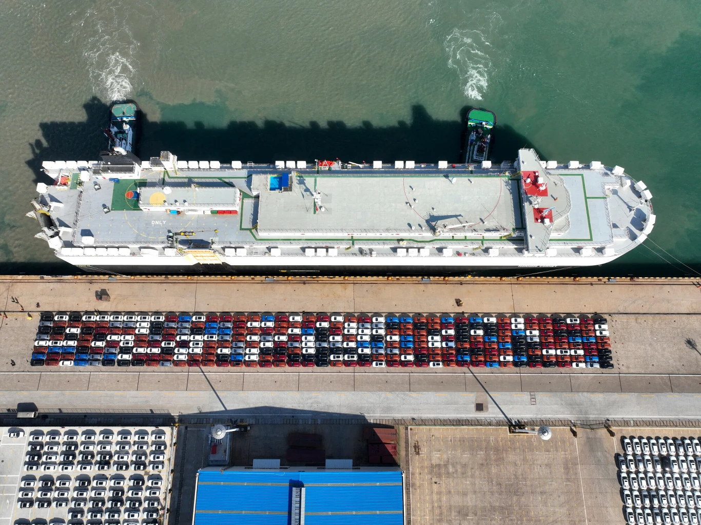 Chiński statek typu ro-ro (zdj. ilustracyjne) Statek transportowy zacumowany przy nabrzeżu, wokół niego zaparkowane setki nowych samochodów w różnych kolorach oczekujących na załadunek lub odbiór, a poniżej fragmenty niebieskiego dachu hali portowej.