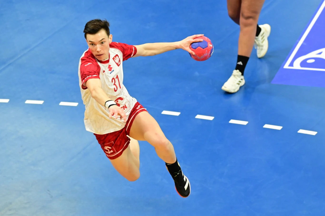 Zawodnik w stroju polskiej drużyny piłki ręcznej wykonuje dynamiczny skok w kierunku bramki trzymając piłkę w prawej ręce, na tle niebieskiego boiska z widocznym fragmentem linii pola bramkowego.