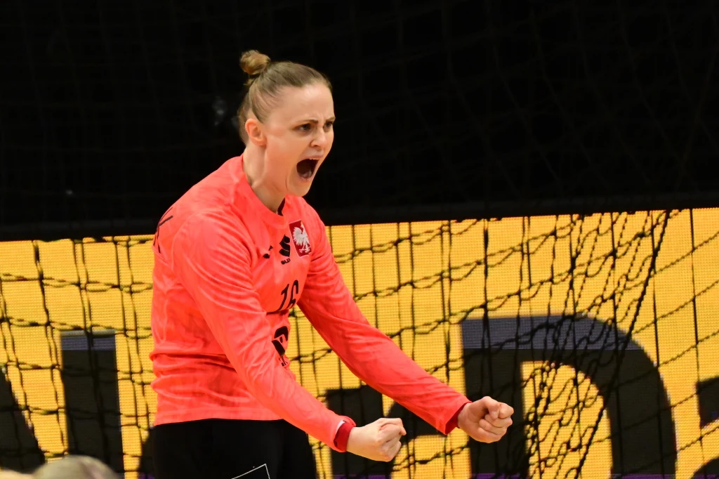 Szczypiornistka w różowym stroju sportowym wyraża emocje i energię stojąc na tle żółtego ekranu i siatki bramki.