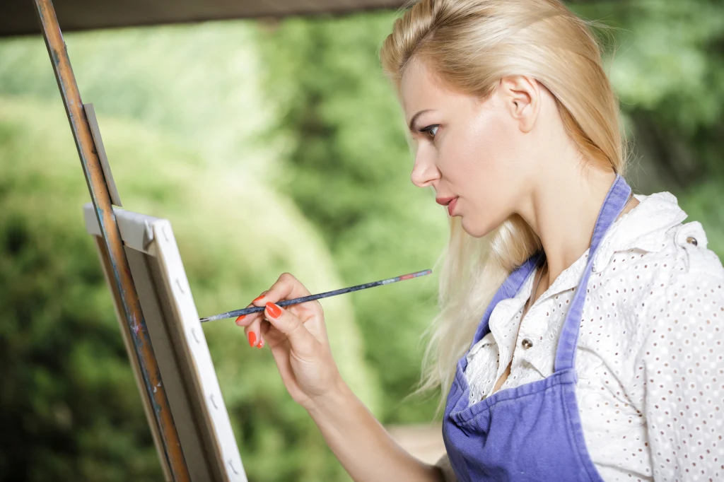 Nie każdy ma talent, by samodzielnie tworzyć obrazy Kobieta blondynka w niebieskim fartuchu skupiona maluje obraz na sztaludze na tle rozmytej zieleni ogrodu lub parku.