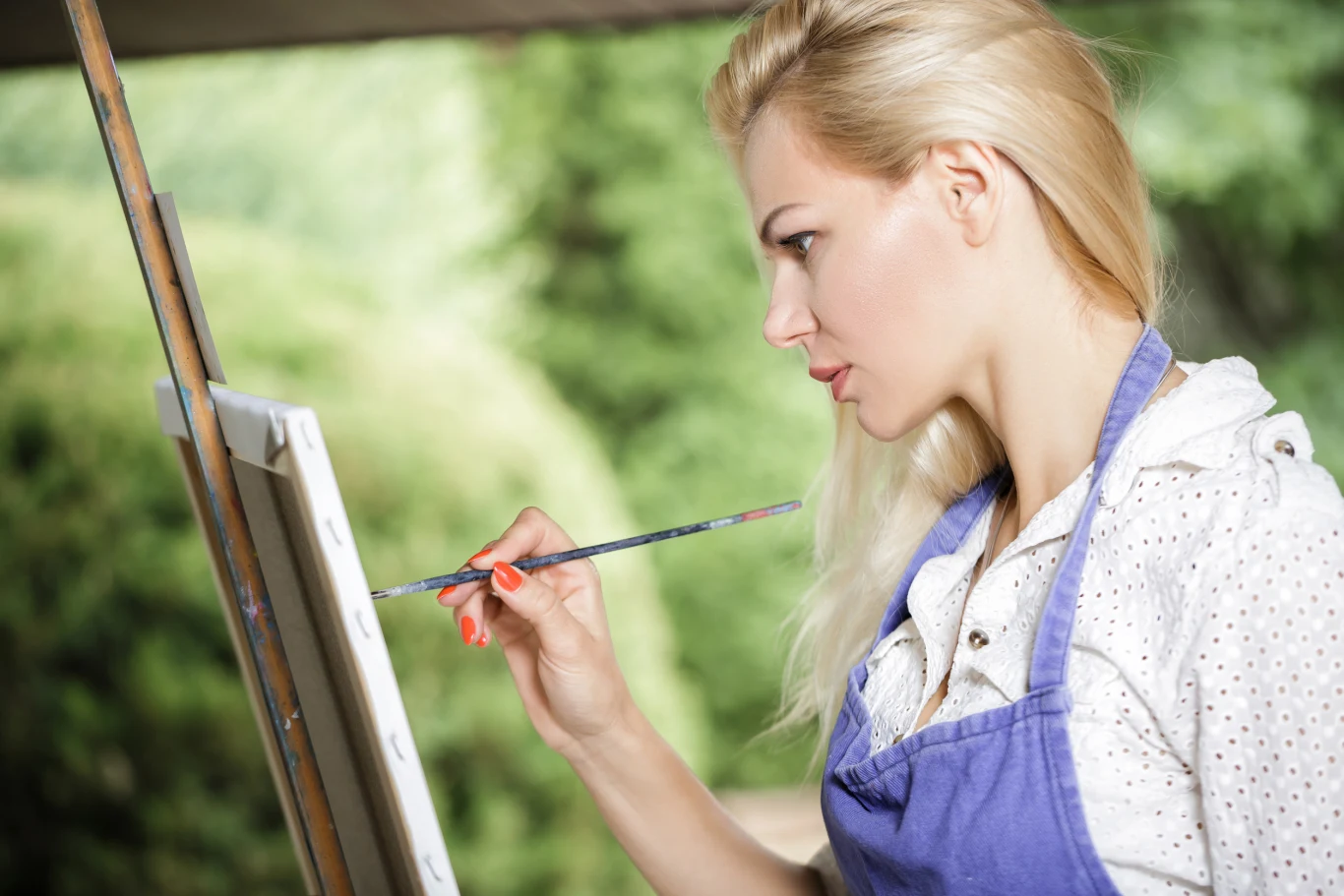 Kobieta blondynka w niebieskim fartuchu skupiona maluje obraz na sztaludze na tle rozmytej zieleni ogrodu lub parku.