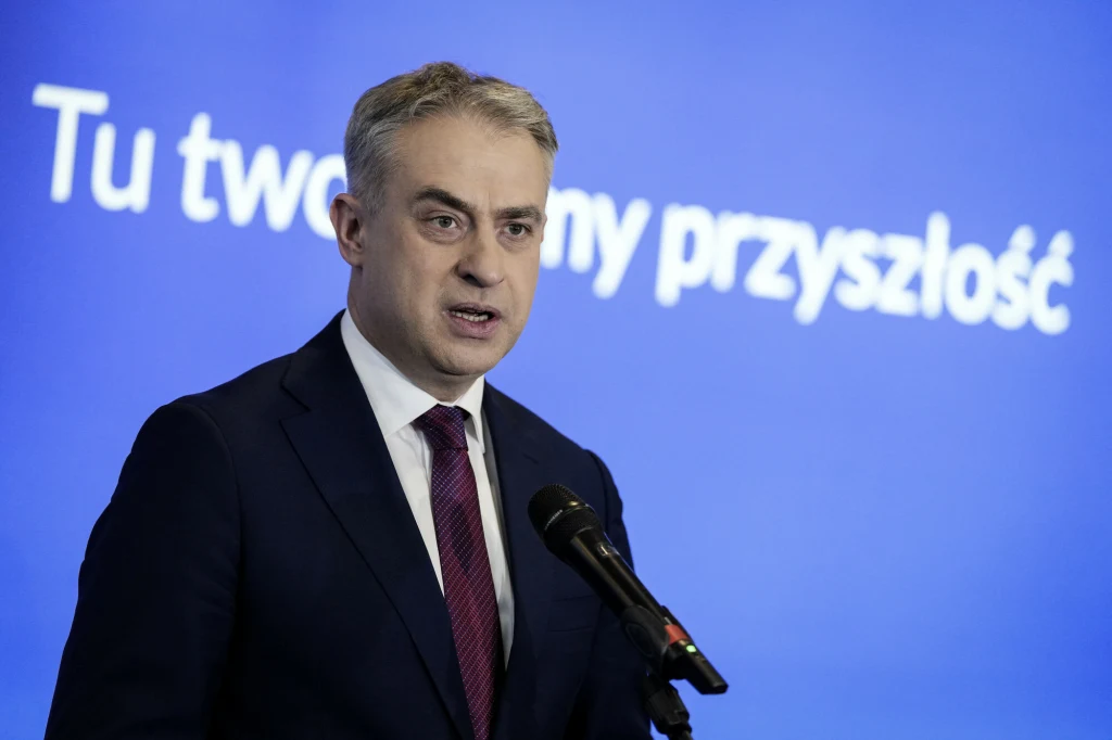 Wicepremier Krzysztof Gawkowski zapewnia, że Nowa Lewica nie dopuści do ograniczenia wydatków na ochronę zdrowia w Polsce Mężczyzna w garniturze i krawacie stoi przy mikrofonie na tle niebieskiego ekranu z białym napisem "Tu tworzymy przyszłość".