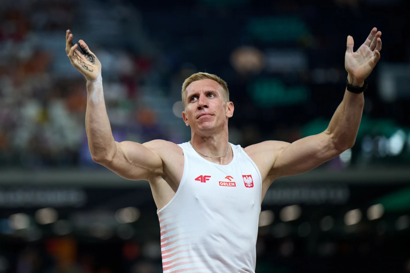 Piotr Lisek Sportowiec w biało-czerwonej koszulce z polskim godłem, prezentuje umięśnione ramiona uniesione do góry, stojąc na stadionie lekkoatletycznym. Twarz skoncentrowana, w tle rozmyte trybuny pełne widzów.