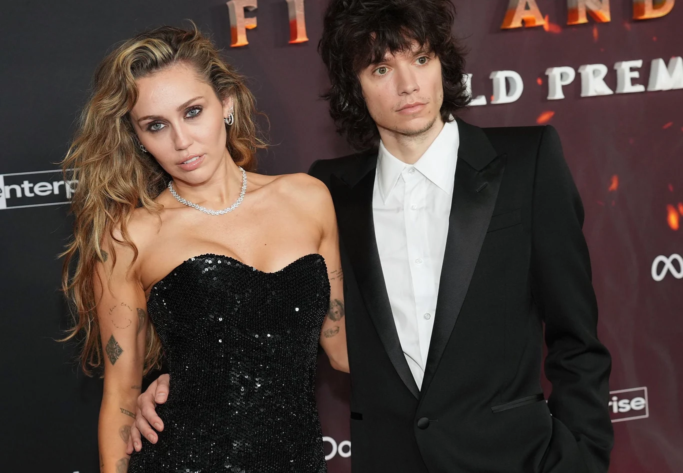 Miley Cyrus i Maxx Morando potwierdzili zaręczyny Kobieta w sukience wieczorowej z cekinami i naszyjnikiem oraz mężczyzna w czarnym smokingu i białej koszuli pozują razem na tle ściany z napisem związanym z premierą filmu lub wydarzenia.