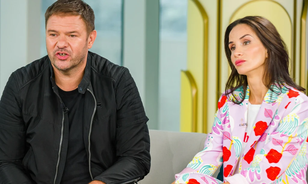 Dwie osoby, mężczyzna ubrany w czarną kurtkę oraz kobieta w kolorowym garniturze ze wzorami kwiatowymi, siedzą obok siebie na sofie w jasnym, nowoczesnym wnętrzu, patrząc w różne kierunki.