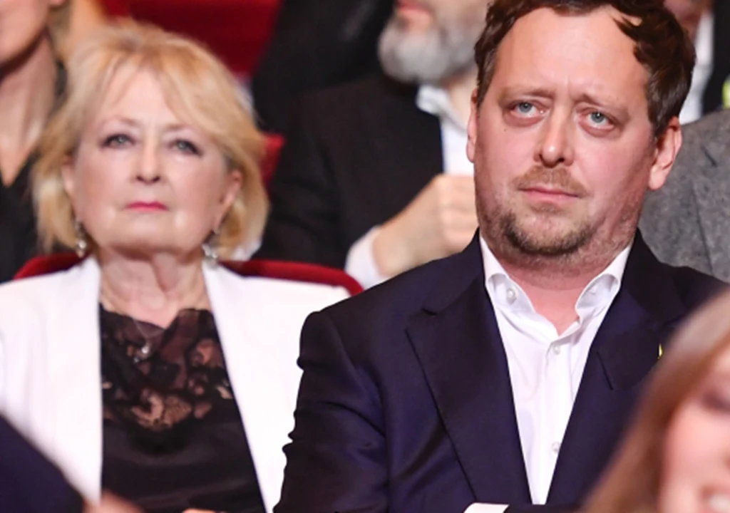 Magdalena Zawadzka i Jan Holoubek Dwie osoby siedzące na publiczności, mężczyzna w ciemnej marynarce z poważnym wyrazem twarzy na pierwszym planie oraz starsza kobieta w jasnej marynarce w tle, rozmyte sylwetki innych osób w tle.