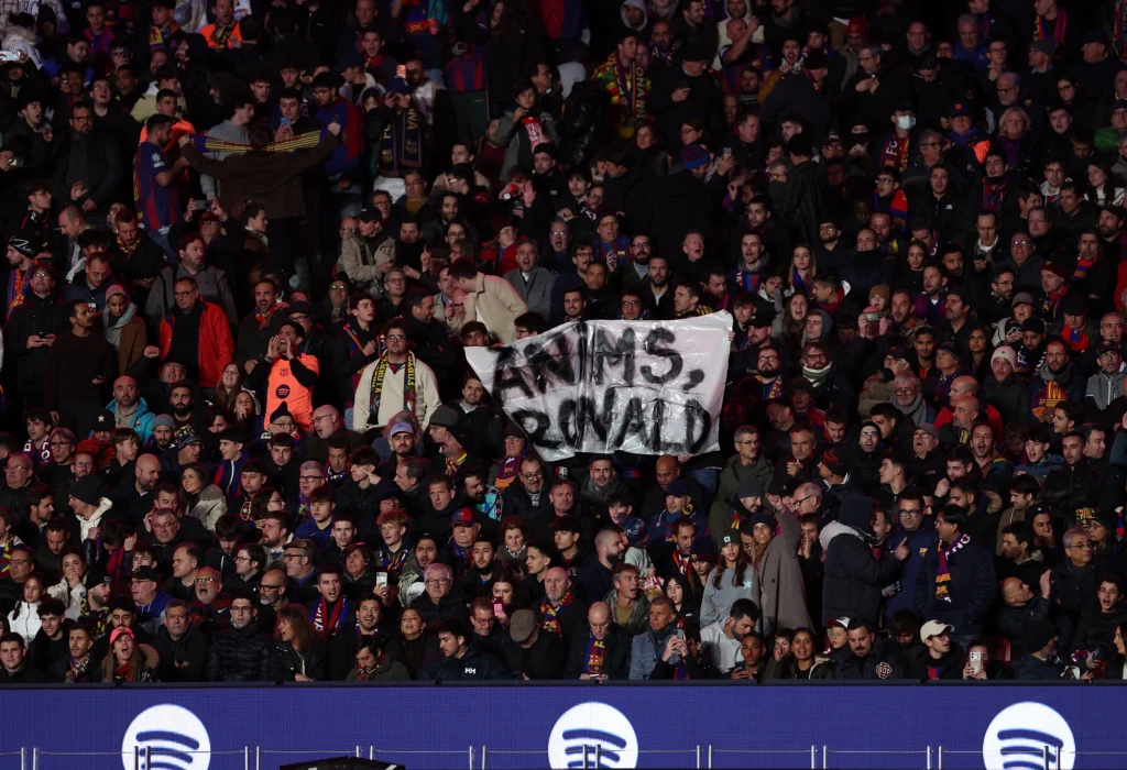 Kibice Barcelony wspierają nieobecnego Ronalda Araujo Tłum kibiców piłkarskich na trybunach stadionu, w centrum grupa osób trzyma transparent z napisem 'Anims Ronald', otoczona przez innych fanów uważnie obserwujących wydarzenia na boisku.