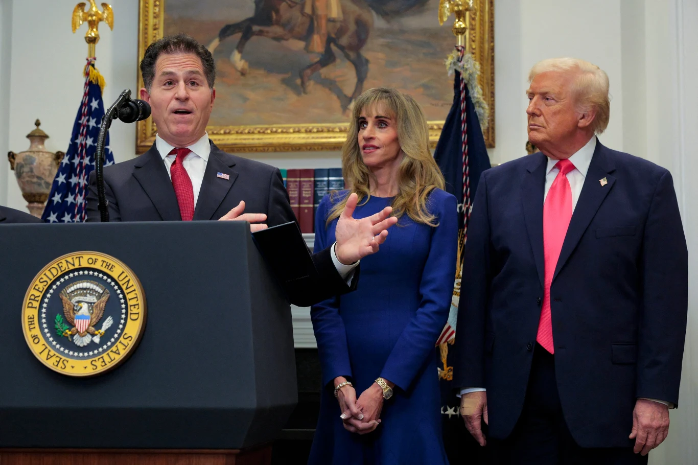 Rodzina Dell przekaże 6 miliardów dolarów na "konta Trumpa" Trzy osoby w eleganckich strojach stoją przy mównicy z prezydenckim godłem Stanów Zjednoczonych, w tle widoczne flagi oraz obraz z jeźdźcem na koniu.