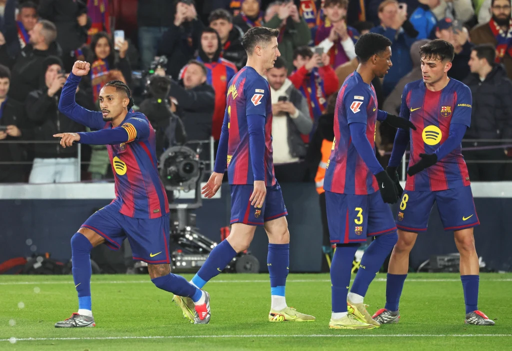 FC Barcelona Piłkarze FC Barcelony w oficjalnych strojach klubowych na boisku podczas meczu piłkarskiego, jeden z nich wykonuje gest po zdobyciu bramki, pozostali zawodnicy zmierzają w jego kierunku, w tle widoczni kibice.