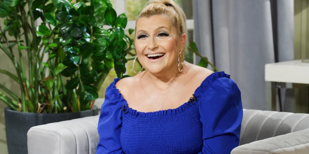 Katarzyna Skrzynecka Kobieta z blond włosami upiętymi w kok, uśmiechnięta, ubrana w niebieską sukienkę z bufiastymi rękawami, siedzi na jasnej sofie, w tle zielona roślina doniczkowa oraz szare zasłony.