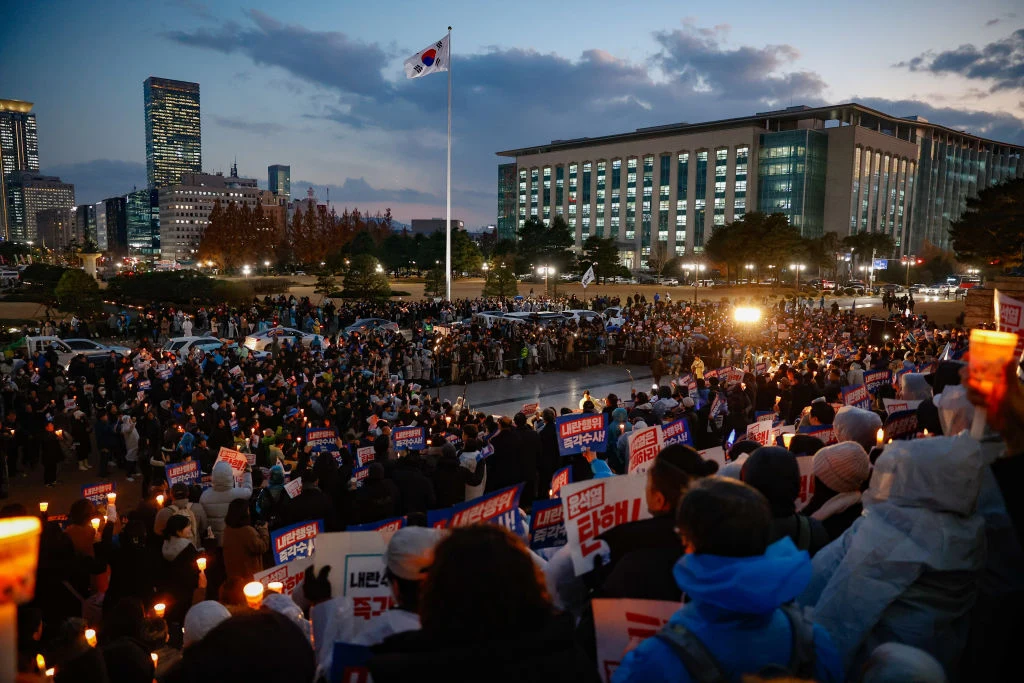 Rocznica wprowadzenia stanu wojennego w Korei Południowej. Na zdjęciu protesty z grudnia 2024 roku w Seulu Tłum ludzi zgromadzony na dużym placu, wielu uczestników trzyma zapalone świece oraz transparenty z napisami, w tle powiewa flaga Korei Południowej, a za tłumem widoczne są nowoczesne budynki podświetlone w czasie zmierzchu.