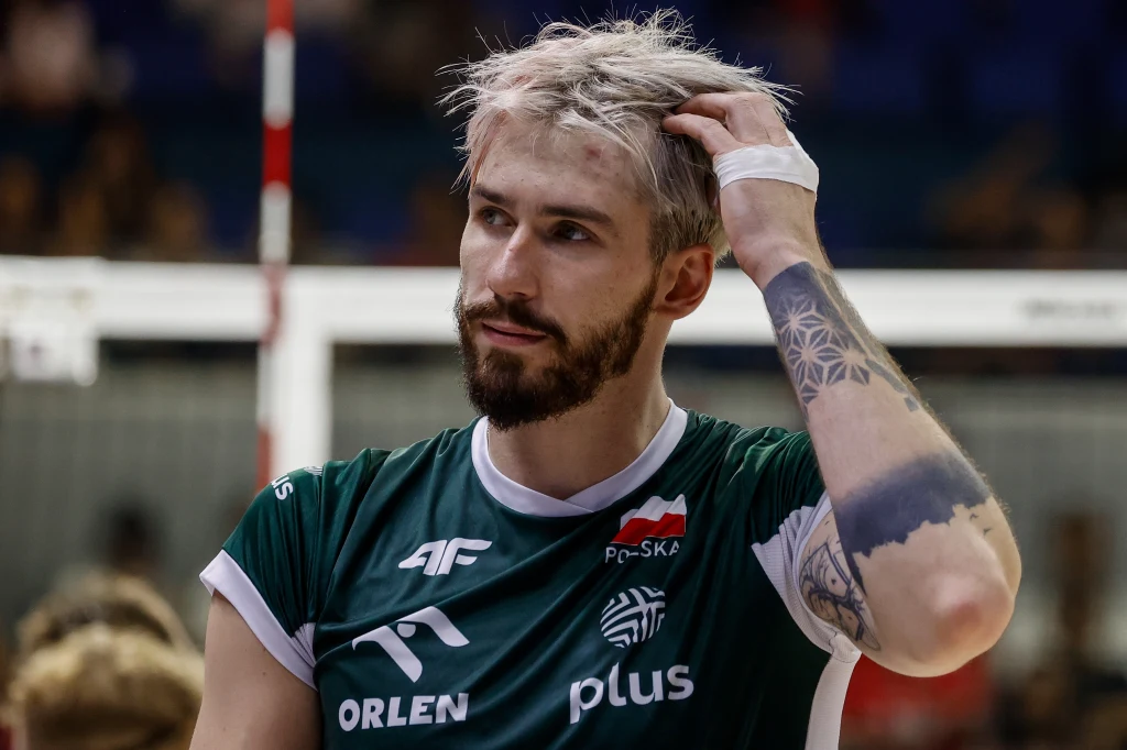 Siatkarz w zielonej koszulce z logo sponsorów i polską flagą na piersi zbiera włosy ręką, widoczny tatuaż na przedramieniu, w tle rozmyte elementy siatki i hali sportowej.