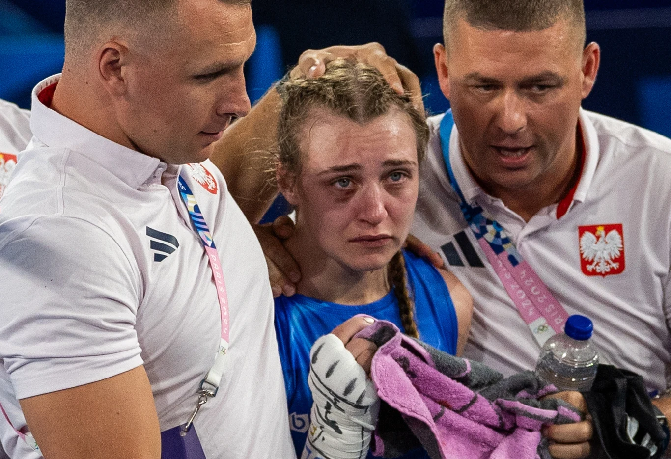 Julia Szeremeta ze sztabem Zmęczona i zapłakana zawodniczka boksu w niebieskim stroju jest pocieszana przez dwóch trenerów w białych koszulkach z polskim godłem. Jeden z nich trzyma ręcznik, drugi butelkę wody.