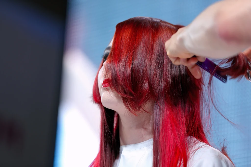 Fryzjer stylizuje intensywnie czerwone włosy kobiety o prostych włosach, skupiając się na precyzyjnym cięciu grzywki. Tło delikatnie rozmyte, skupienie na włosach i dłoniach stylizującego.