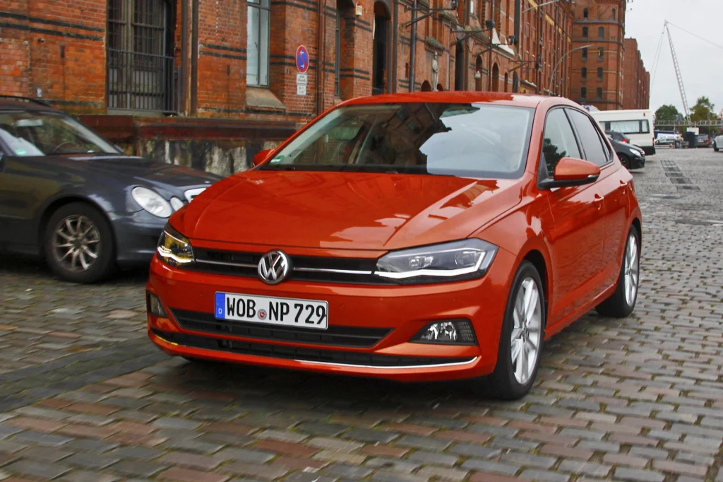 Czerwony samochód osobowy marki Volkswagen Polo zaparkowany na brukowanej ulicy miejskiej, w tle widoczne ceglane budynki oraz inne pojazdy.