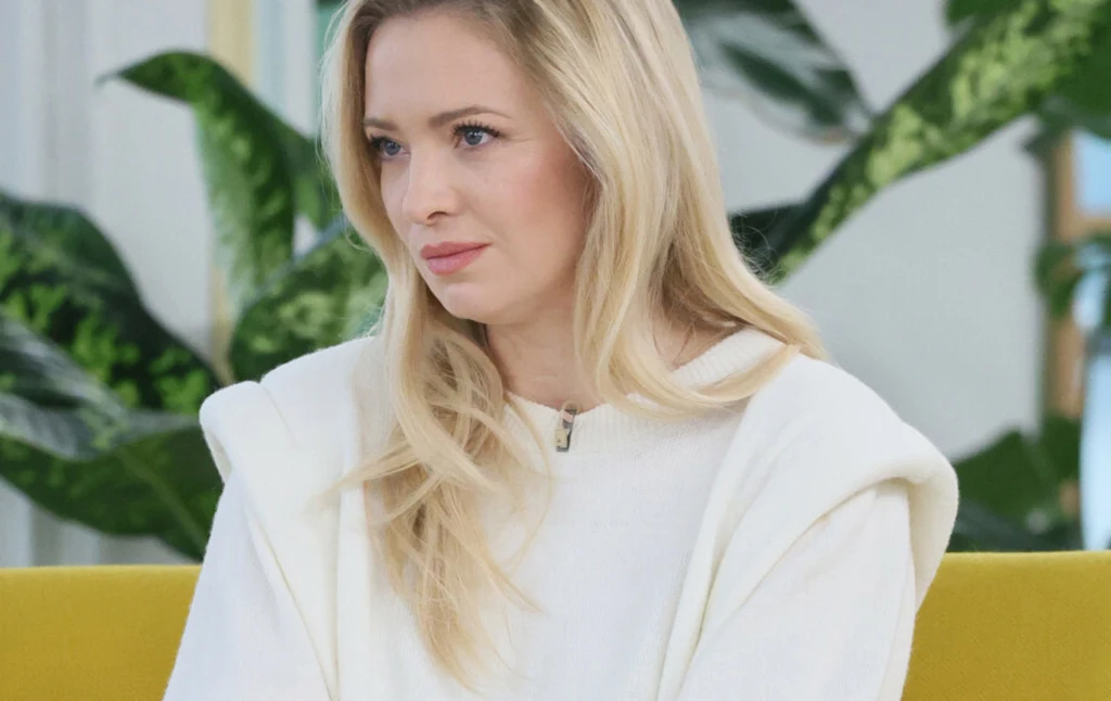 Kobieta z blond włosami ubrana w jasną bluzę siedzi na żółtej sofie, w tle widoczne są zielone rośliny ozdobne.