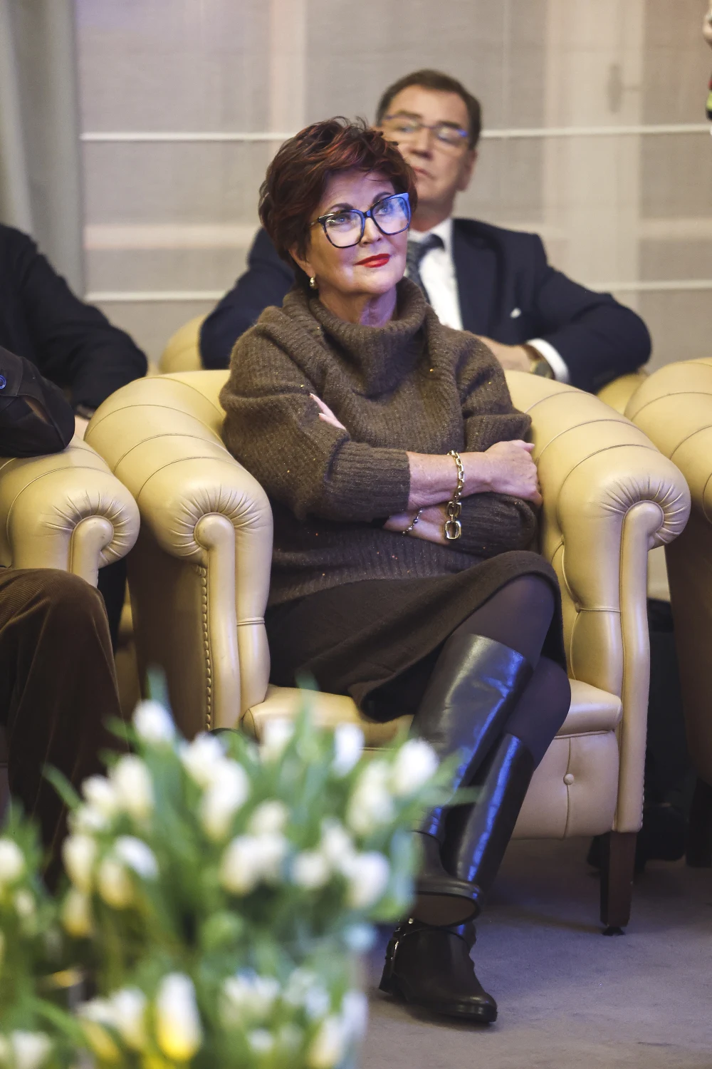 Jolanta Kwaśniewska na spotkaniu promującym autobiografię „Mariusz Walter. Miłość i telewizja” Kobieta w okularach i ciemnym swetrze siedzi na jasnym, skórzanym fotelu z założonymi rękami, w tle siedzi mężczyzna w garniturze, z przodu widoczny bukiet tulipanów.