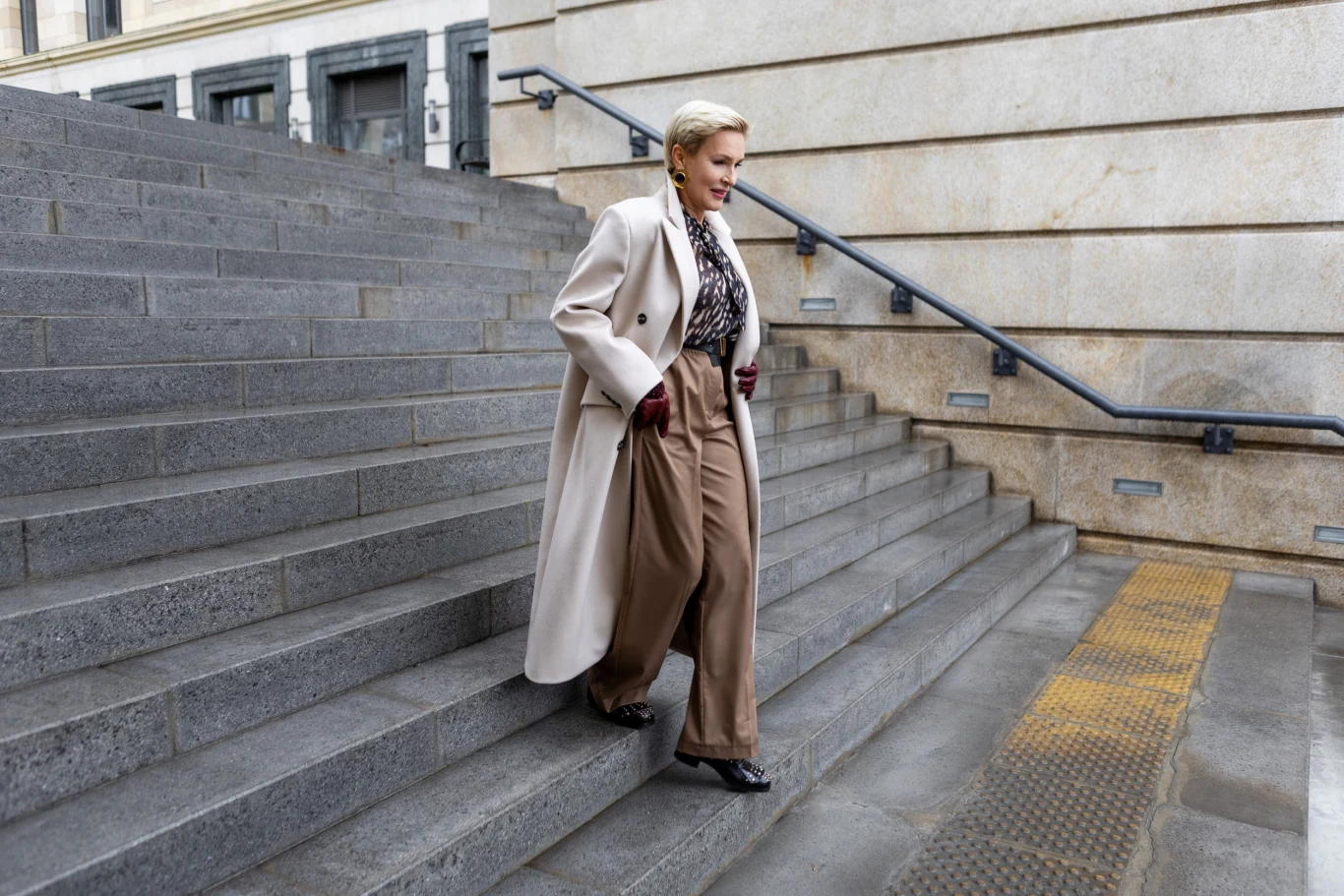 Elegancko ubrana kobieta schodzi po szerokich, kamiennych schodach miejskich. Ma na sobie beżowy płaszcz, luźne spodnie i ciemne buty. W tle widoczna jest fasada budynku z kamienia.