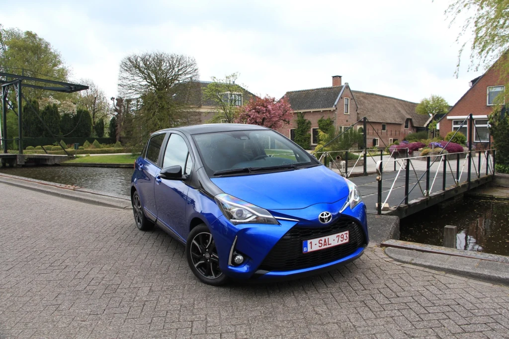 Niebieski samochód osobowy Toyota Yaris zaparkowany przy brukowanej ulicy w spokojnej, wiejskiej okolicy z charakterystycznymi domami i mostkiem nad kanałem w tle.