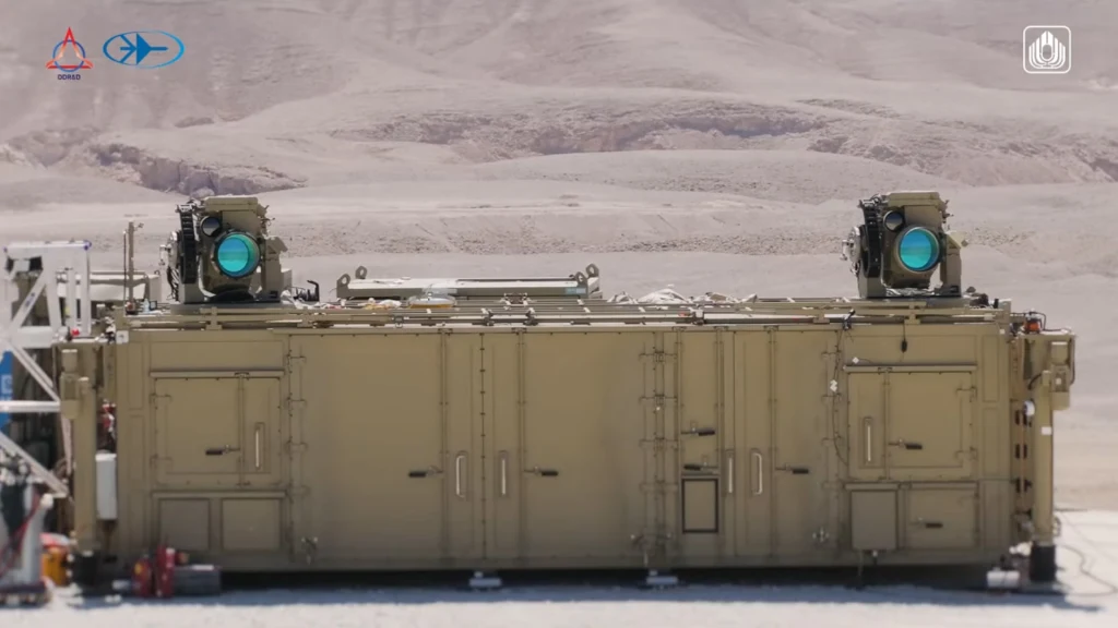 Duży pancerny moduł techniczny ustawiony na pustynnym tle, na dachu zamontowane dwie duże wieżyczki z widocznymi soczewkami laserowymi koloru niebieskiego. Konstrukcja jednolita, prostokątna, wykonana w piaskowym kolorze dopasowanym do otoczenia. Brak ...