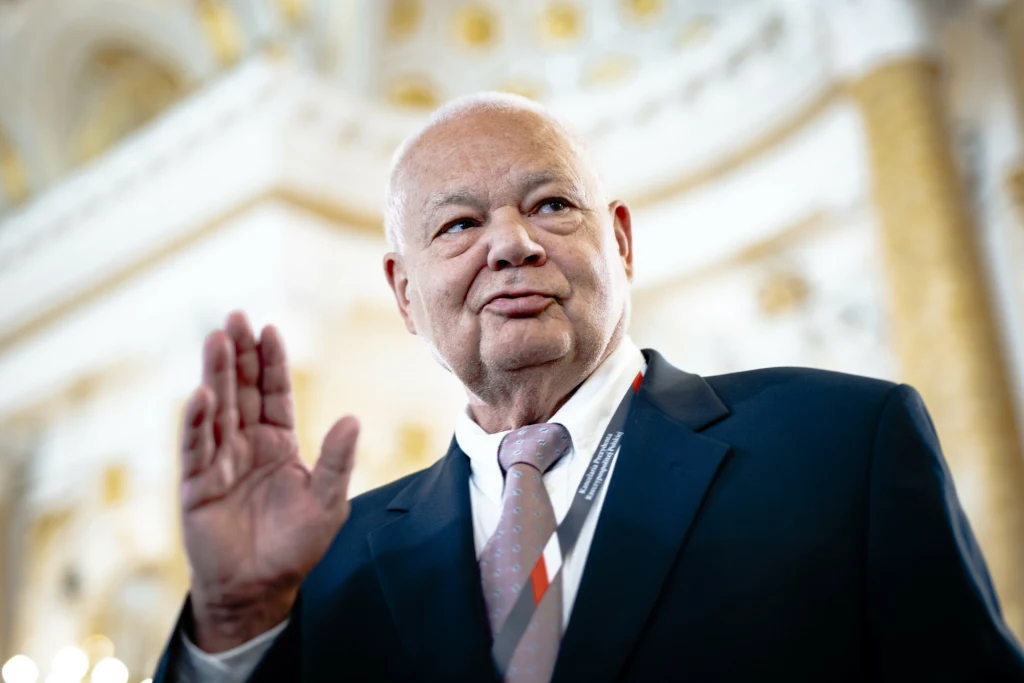 Starszy, łysy mężczyzna w garniturze i krawacie stoi w jasnym, eleganckim wnętrzu z podniesioną ręką, jakby pozdrawiał lub wygłaszał przemówienie. Za nim widać rozmyte architektoniczne detale, sugerujące oficjalne miejsce.