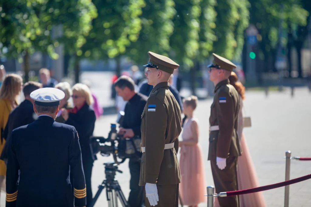 Dwóch żołnierzy w uniformach stoi na tle grupy ludzi, w tym kamerzystów i gości, w miejscu publicznym otoczonym zielenią, z elementami barierek sugerujących oficjalny charakter wydarzenia.