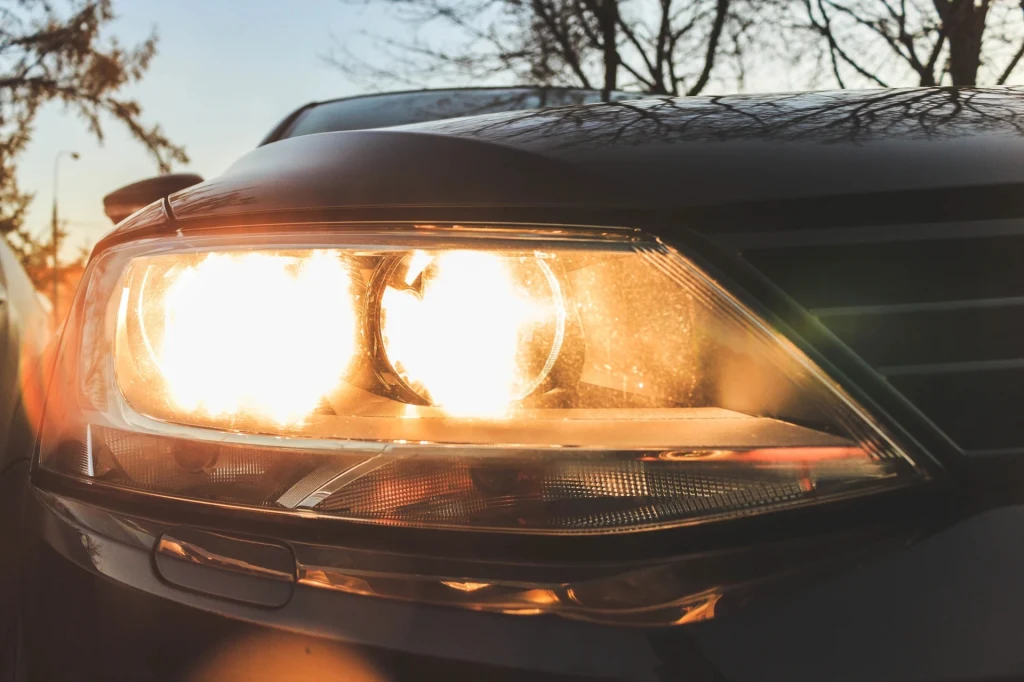 Reflektor samochodowy włączony podczas zachodu słońca, światło odbija się na powierzchni karoserii, w tle widoczne nagie gałęzie drzew.
