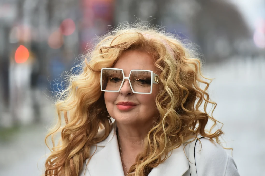 Magda Gessler ma na to sposób. Jej dodatki robią cały efekt Kobieta o długich, kręconych blond włosach, ubrana w jasny płaszcz i duże, kwadratowe okulary, stoi na nieostrym tle miejskiej ulicy.