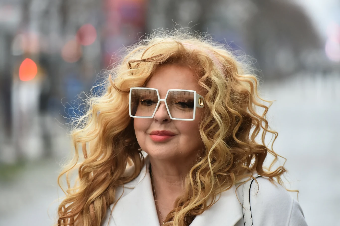 Magda Gessler ma na to sposób. Jej dodatki robią cały efekt Kobieta o długich, kręconych blond włosach, ubrana w jasny płaszcz i duże, kwadratowe okulary, stoi na nieostrym tle miejskiej ulicy.