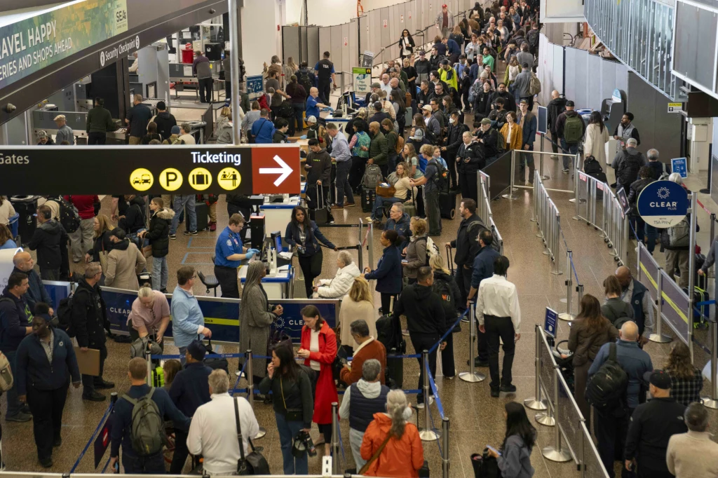 Lotnisko w USA. Administracja Donalda Trumpa wstrzymuje procedowanie wniosków migracyjnych dla 19 państw Długie kolejki ludzi czekających na odprawę na lotnisku, tłum pasażerów z bagażami, oddzielone taśmami stanowiska, oznaczenia kierunkowe i tablice informacyjne nad głowami.