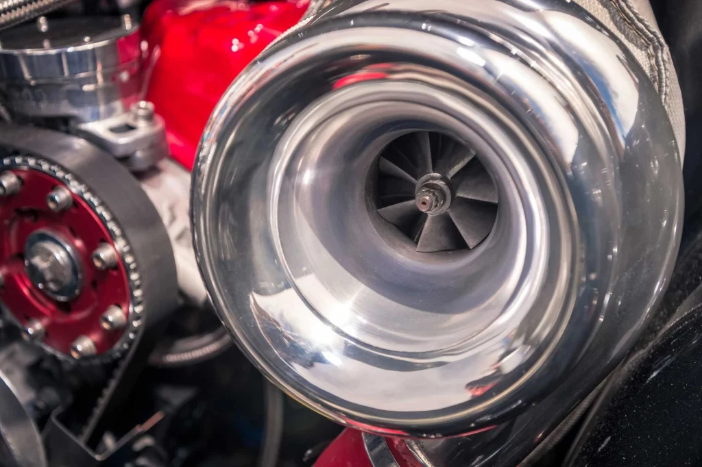 Zbliżenie na połyskującą turbosprężarkę zamontowaną w komorze silnika, widoczne detale takie jak łopatki wirnika, metalowe elementy oraz fragment czerwonego korpusu mechanicznego.