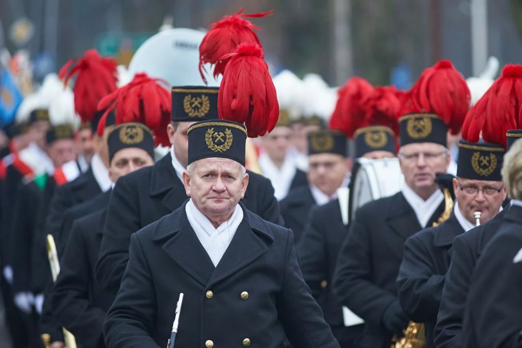 Barbórka to nie tylko parady, ale również wypłaty nagród dla górników Górnicy w tradycyjnych mundurach z czarnymi czapkami ozdobionymi czerwonymi pióropuszami idą w zwartym pochodzie, wyraziste emblematy i poważne twarze podkreślają uroczysty charakter wydarzenia.