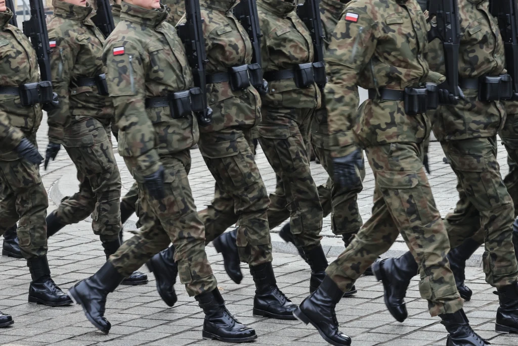 Żołnierze w mundurach wojskowych maszerują w szyku podczas parady wojskowej, trzymając broń przewieszoną przez ramię. Na mundurach widoczne są polskie flagi.