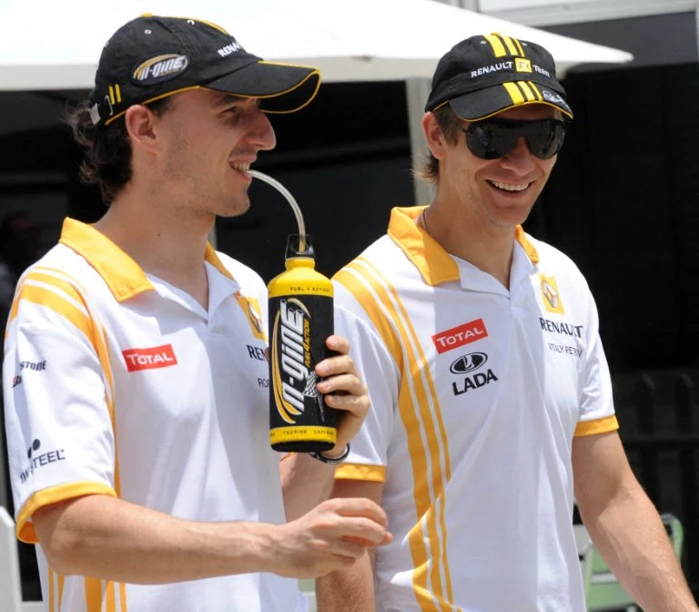 Dwóch mężczyzn w sportowych koszulkach zespołu Renault F1 uśmiecha się idąc razem, jeden z nich trzyma bidon z napojem i pije przez rurkę. Obaj mają czapki z daszkiem i okulary przeciwsłoneczne.