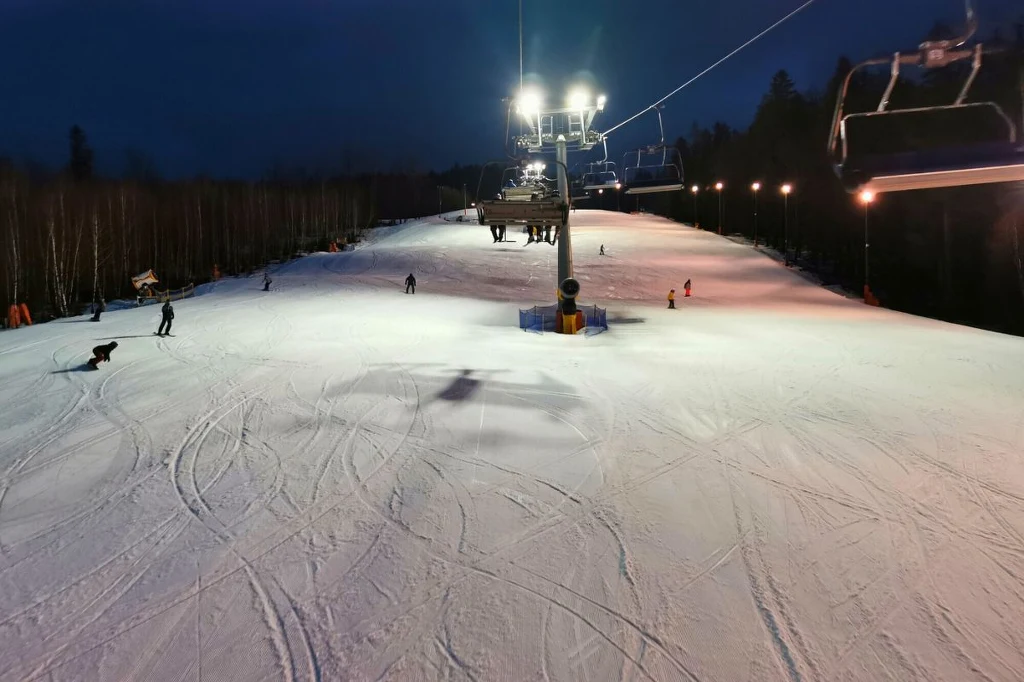 Oświetlony stok narciarski z kilkoma osobami jeżdżącymi na nartach i snowboardzie, po prawej stronie widoczna czynna kolejka krzesełkowa, wokół las iglasty, a w tle ciemne niebo.