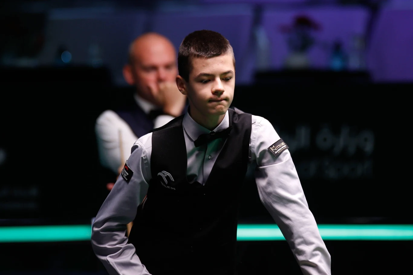 Młody zawodnik snookera w kamizelce i muszce stoi przy stole, z poważnym wyrazem twarzy, w tle widać innego mężczyznę skupionego na grze.