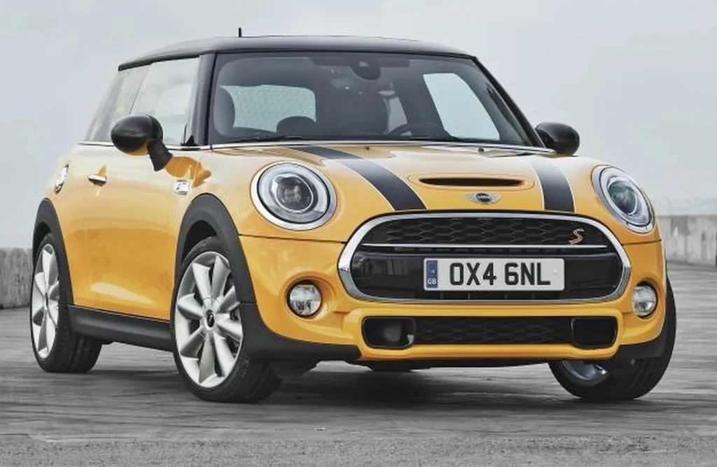 Żółty samochód Mini Cooper z czarnymi pasami na masce stoi na utwardzonej powierzchni, z widoczną tablicą rejestracyjną i srebrnymi felgami, tło stanowi lekko zamglone niebo.