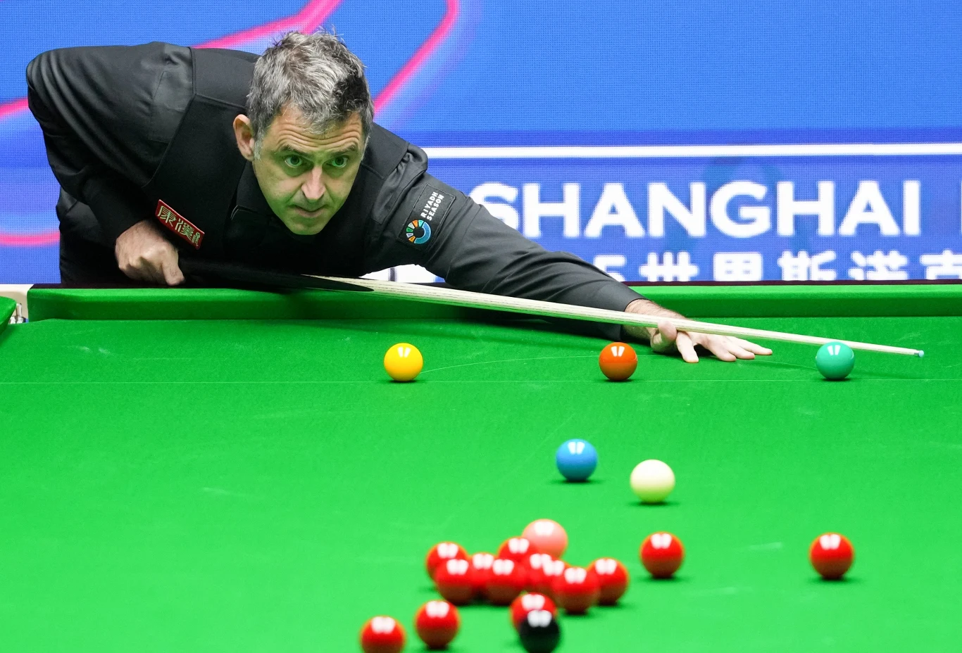 Zawodnik snookera koncentrujący się na wykonaniu uderzenia, wykonuje precyzyjny ruch kijem nad zielonym stołem bilardowym, wokół znajdują się kolorowe bile, w tle widoczny fragment napisu i niebieskie tło z nazwą miasta.