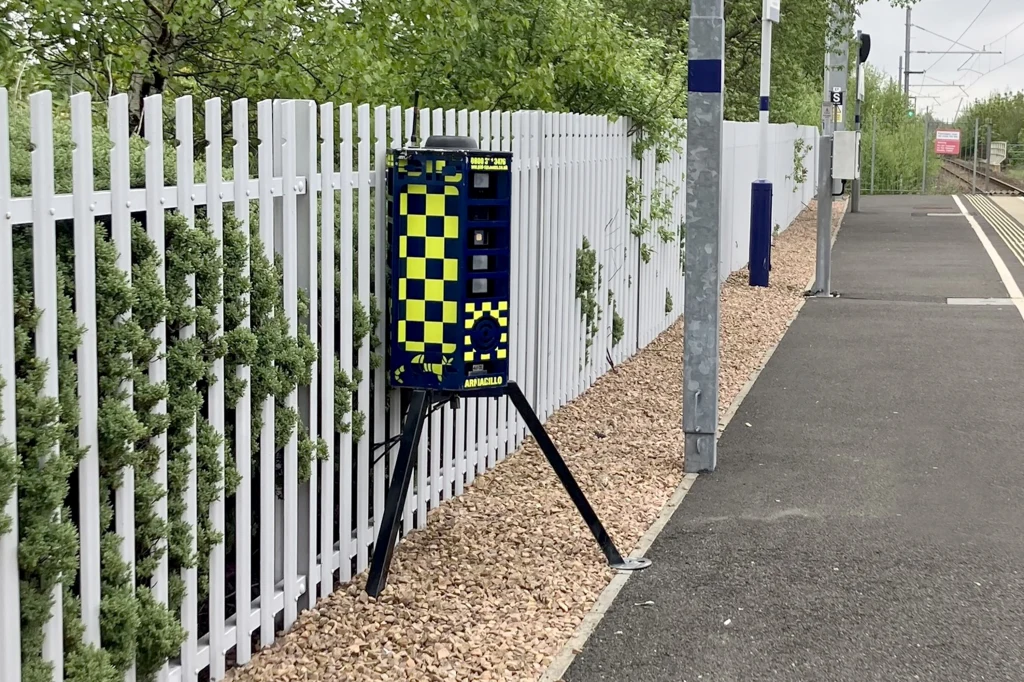 Kierowcy myślą, że to fotoradar i zdejmują nogę z gazu. Fot. DeterTech @Instagram Urządzenie umieszczone na trójnogu przy ogrodzeniu stacji kolejowej, biały metalowy płot i fragment peronu wyłożony asfaltem, w tle tory kolejowe oraz zielone krzewy.