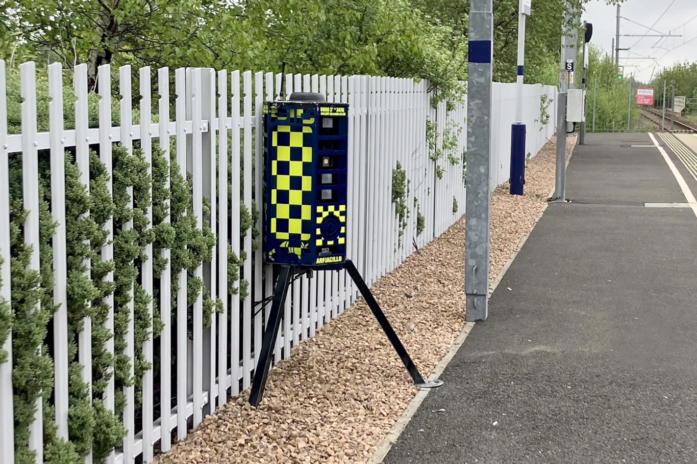 Kierowcy myślą, że to fotoradar i zdejmują nogę z gazu. Fot. DeterTech @Instagram Urządzenie umieszczone na trójnogu przy ogrodzeniu stacji kolejowej, biały metalowy płot i fragment peronu wyłożony asfaltem, w tle tory kolejowe oraz zielone krzewy.