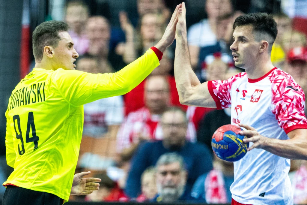 Adam Morawski (z lewej) zakończył reprezentacyjną karierę, Szymon Sićko ją zawiesił. Na EURO obu nie będzie Dwóch polskich piłkarzy ręcznych daje sobie piątkę podczas meczu, jeden z nich ubrany w żółty strój bramkarski z nazwiskiem Morawski i numerem 94, drugi trzyma piłkę do gry, w tle widoczni kibice.