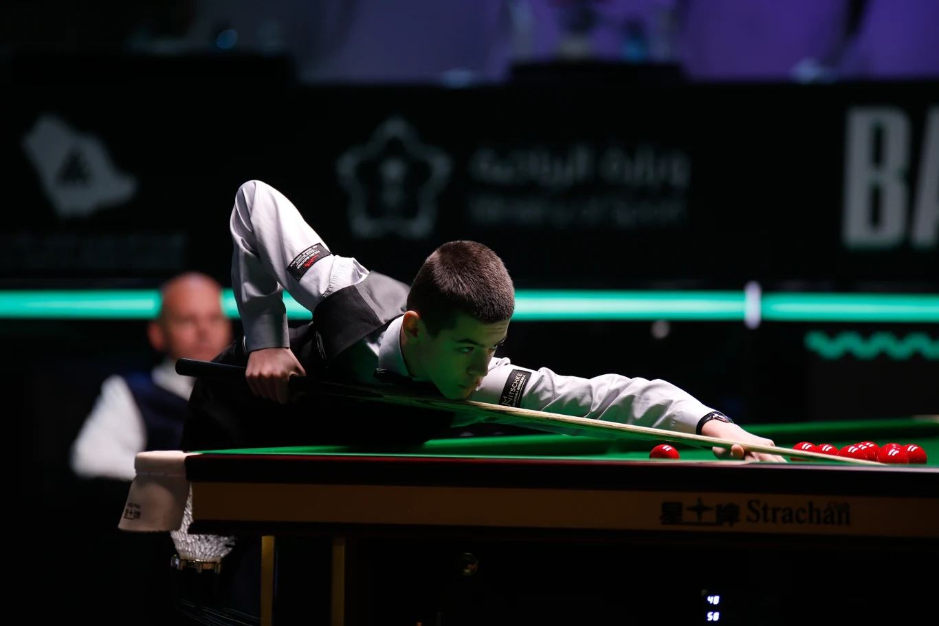 Michał Szubarczyk Młody zawodnik w białej koszuli i kamizelce wykonuje uderzenie w snookerze, skupiając się na precyzyjnym ustawieniu kija tuż nad stołem, przy którym widoczne są czerwone bile. W tle siedzi sędzia lub inny uczestnik meczu.