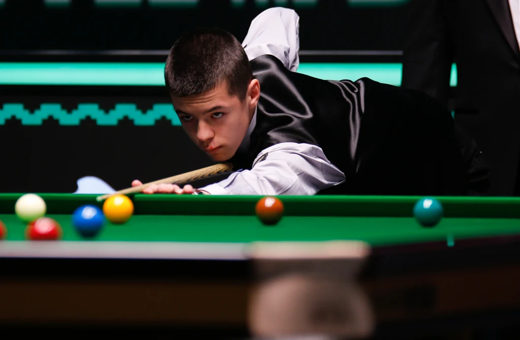 Michał Szubarczyk Młody mężczyzna w eleganckim stroju koncentruje się podczas gry w snookera, trzyma kij bilardowy i celuje do jednej z kolorowych bil.