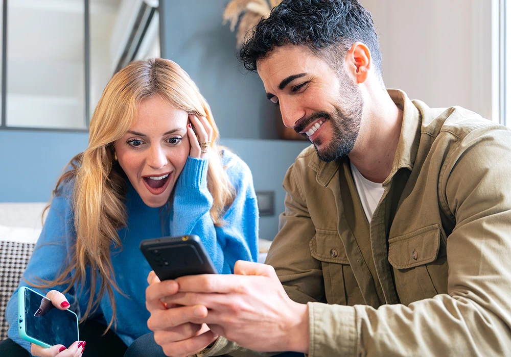Oferty „bez limitu” są idealne dla osób, które korzystają z internetu intensywnie – oglądają filmy, pracują zdalnie, grają lub prowadzą transmisje Młoda kobieta i mężczyzna siedzą razem na kanapie, oboje trzymają telefony. Kobieta wyraża zaskoczenie i radość, a mężczyzna z uśmiechem pokazuje jej coś na ekranie swojego telefonu.