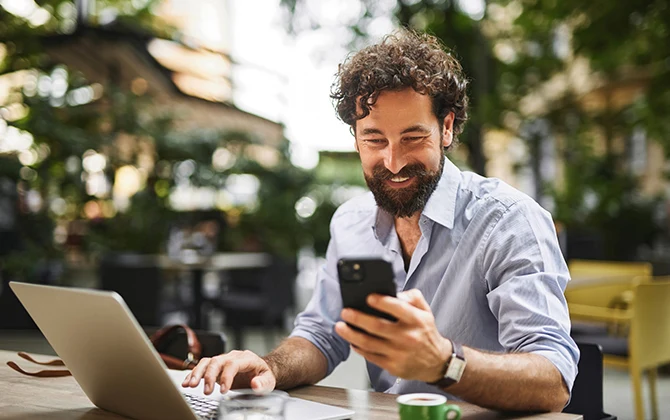 Oszczędzanie na internecie mobilnym wcale nie musi oznaczać rezygnacji z wygody Mężczyzna z brodą siedzi przy stole na zewnątrz, trzyma smartfon i uśmiecha się, obok niego laptop, a w tle rozmyte zielone rośliny.