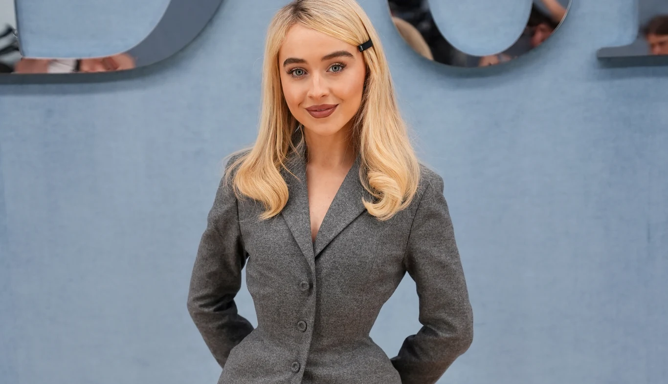 Sabrina Carpenter kontra Donald Trump Młoda kobieta o blond włosach z grzywką spiętą spinką, ubrana w elegancki, szary garnitur, stoi na tle jasnoniebieskiej ściany z widoczną literą napisu w tle. Kobieta uśmiecha się i ma dłonie za plecami.