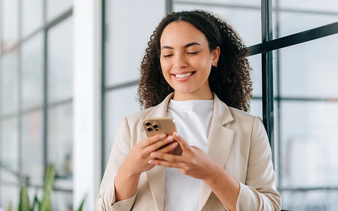 Promocje sezonowe to świetny moment, żeby realnie obniżyć rachunki Kobieta w jasnej marynarce i białej bluzce stoi w nowoczesnym, jasnym biurze, trzyma w rękach smartfon i uśmiecha się patrząc na ekran.